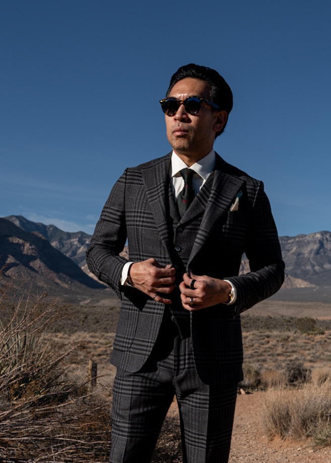 Model wearing the Onyx Charcoal Prince of Wales three-piece suit with sunglasses, standing outdoors in a desert mountain landscape